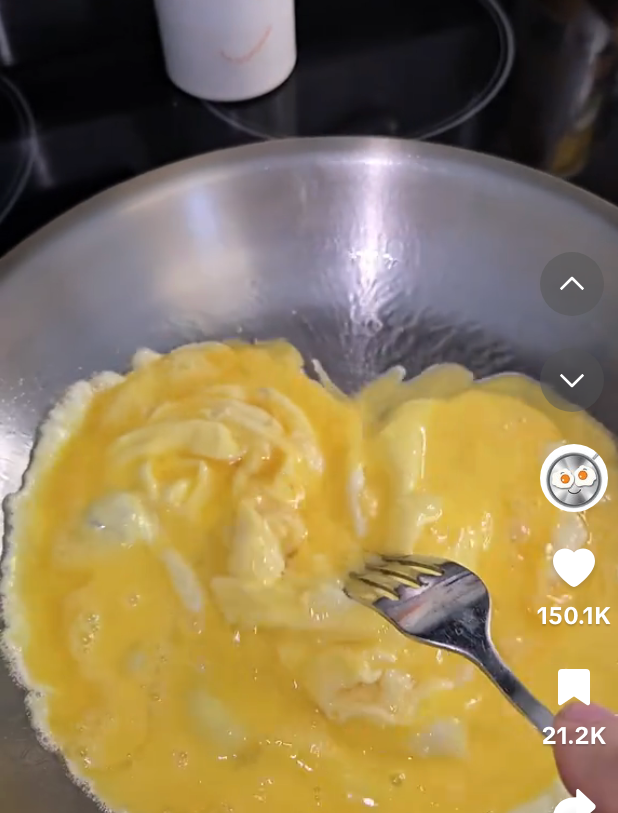 Scrambled eggs being cooked in a stainless steel pan with a fork, as part of a cooking video tutorial