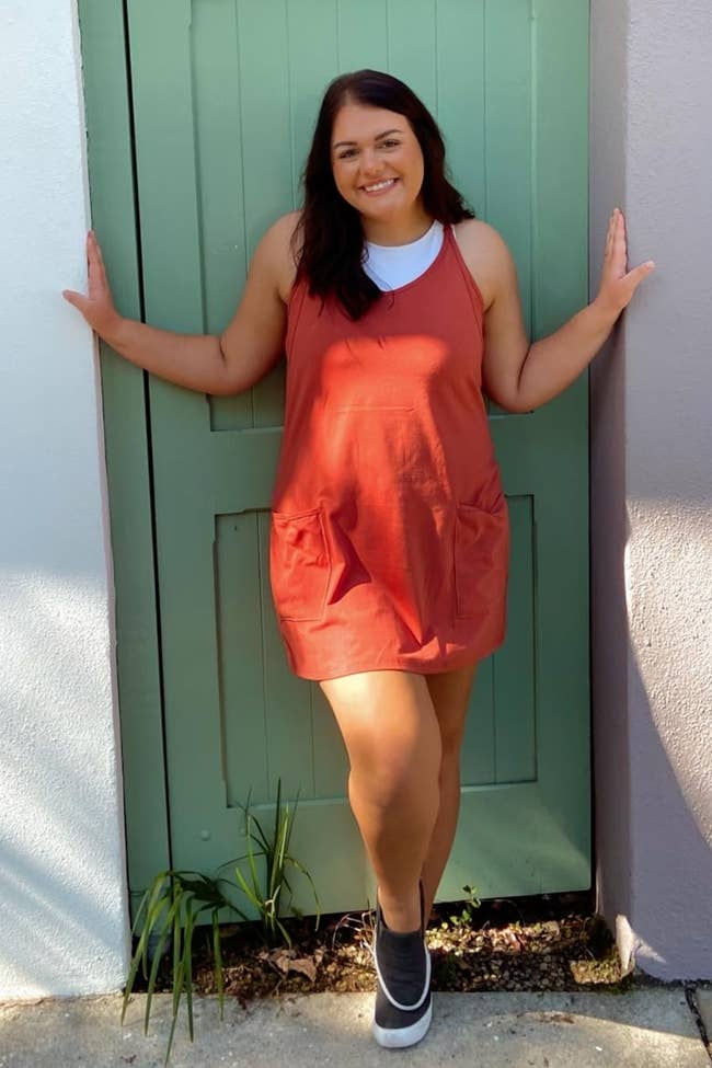Person successful a casual orangish formal stands smiling betwixt colorful walls and gate, wearing sneakers; the nonfiction focuses connected shopping