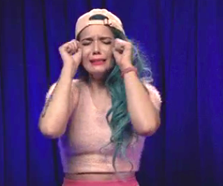 halsey with long hair and cap mimics crying by rubbing eyes comically, wearing a fuzzy top, in a playful or humorous act