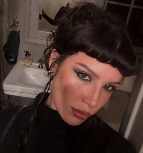 halsey with styled hair takes a selfie in front of a bathroom sink. They wear bold eyeliner and lipstick