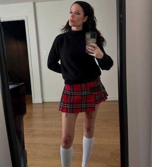 halsey taking a mirror selfie wearing a black sweater and a plaid skirt, with knee-high white socks, standing on a wooden floor