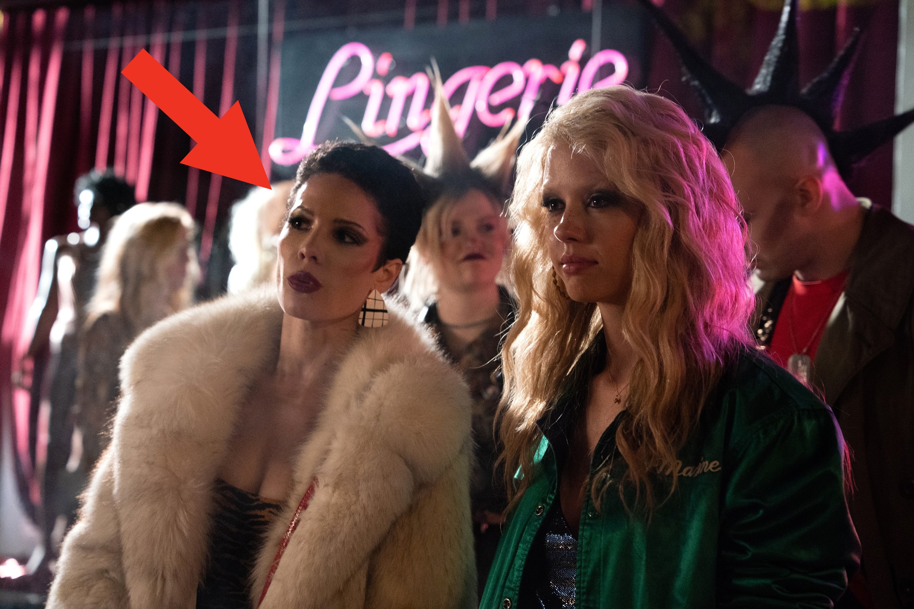 halsey and mia goth stand in a dimly lit room with neon signs One wears a fur coat; the other has a casual jacket People and various hairstyles are in the background