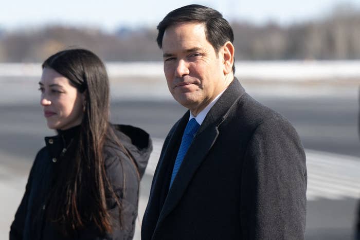 A man in a suit and tie stands next to a woman in a coat at an outdoor event, looking toward the camera