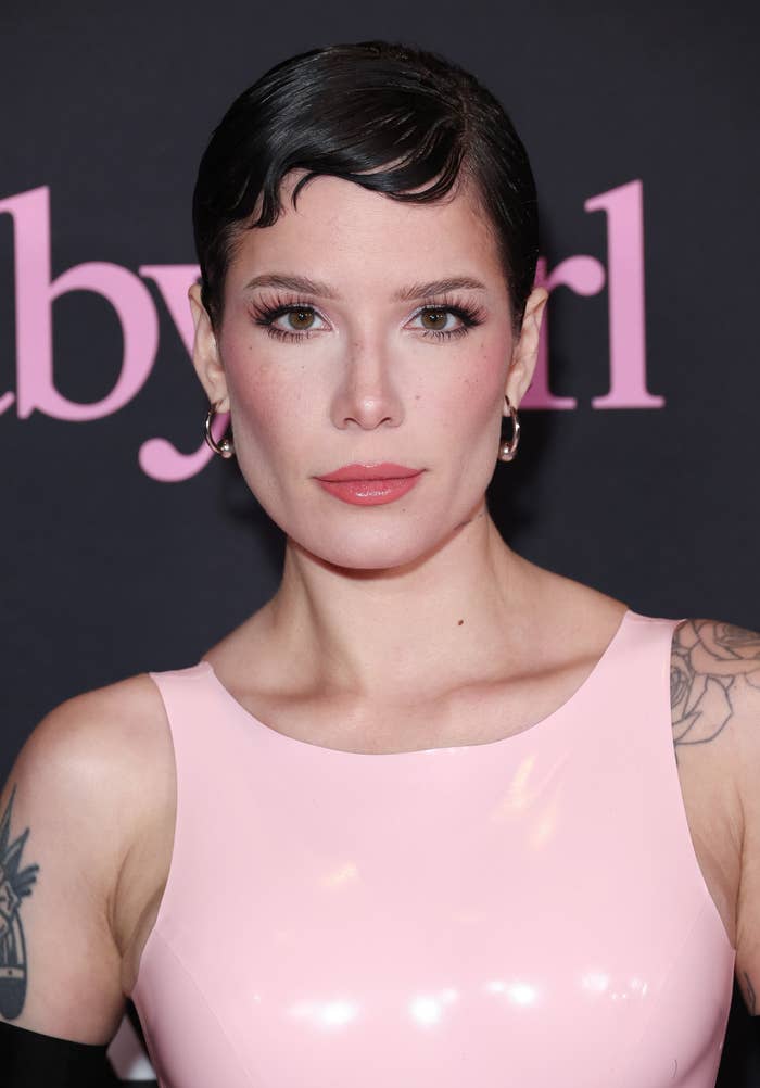 halsey in a glossy outfit with geometric haircut, hoop earrings, and visible tattoos standing against a backdrop with partial text