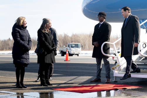 A group of people stand near an airplane with one person on the stairs. The setting appears formal, with a red carpet on the ground