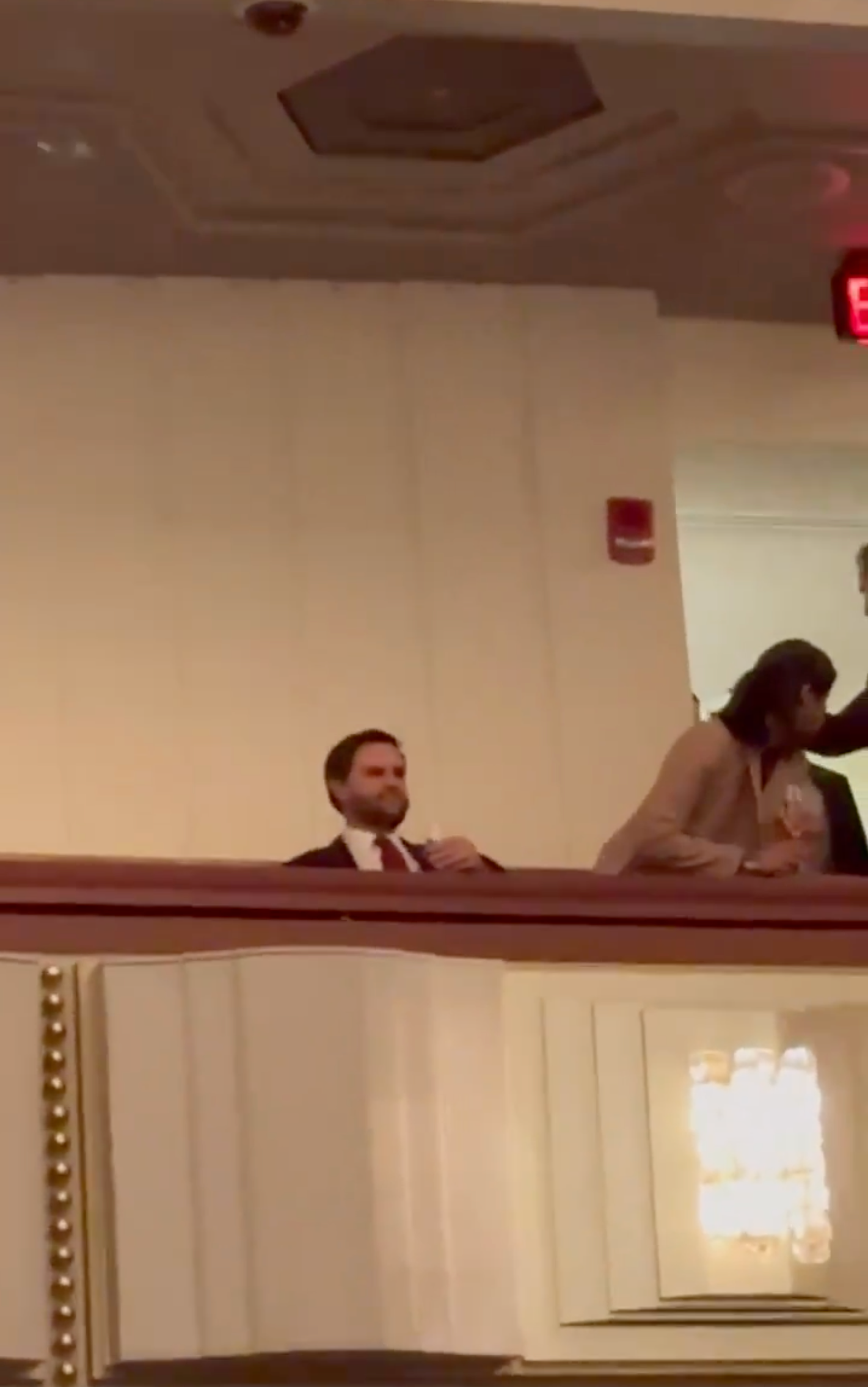 Two people in a theater balcony; one in a dark suit seated, the other in a light suit standing and talking