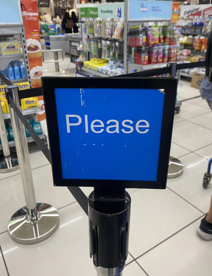 A sign in a store aisle reads &quot;Please,&quot; attached to a pole, likely indicating instructions for customers or marking a queue area