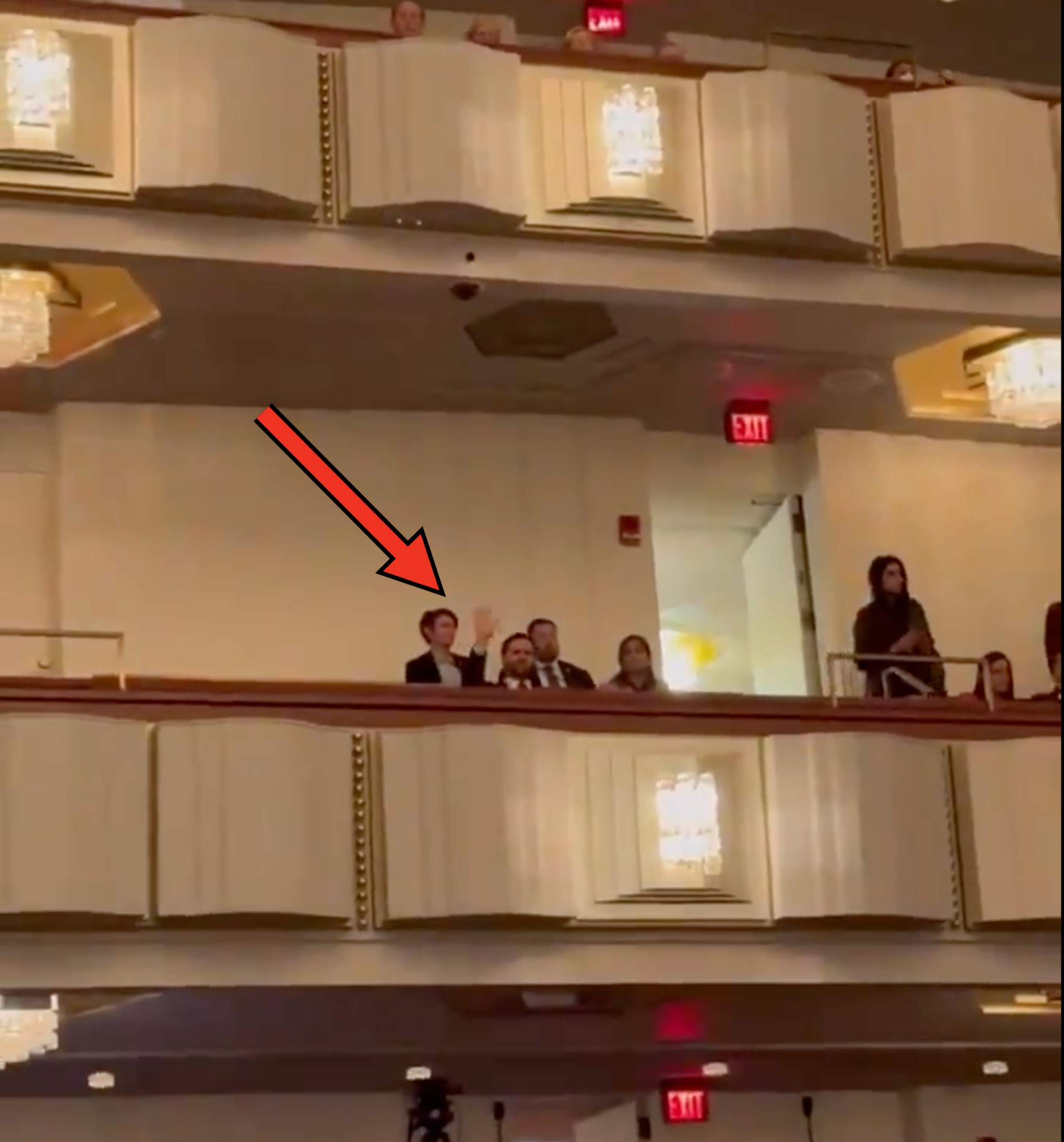 People seated in theater balconies, some waving. Chandelier lighting visible