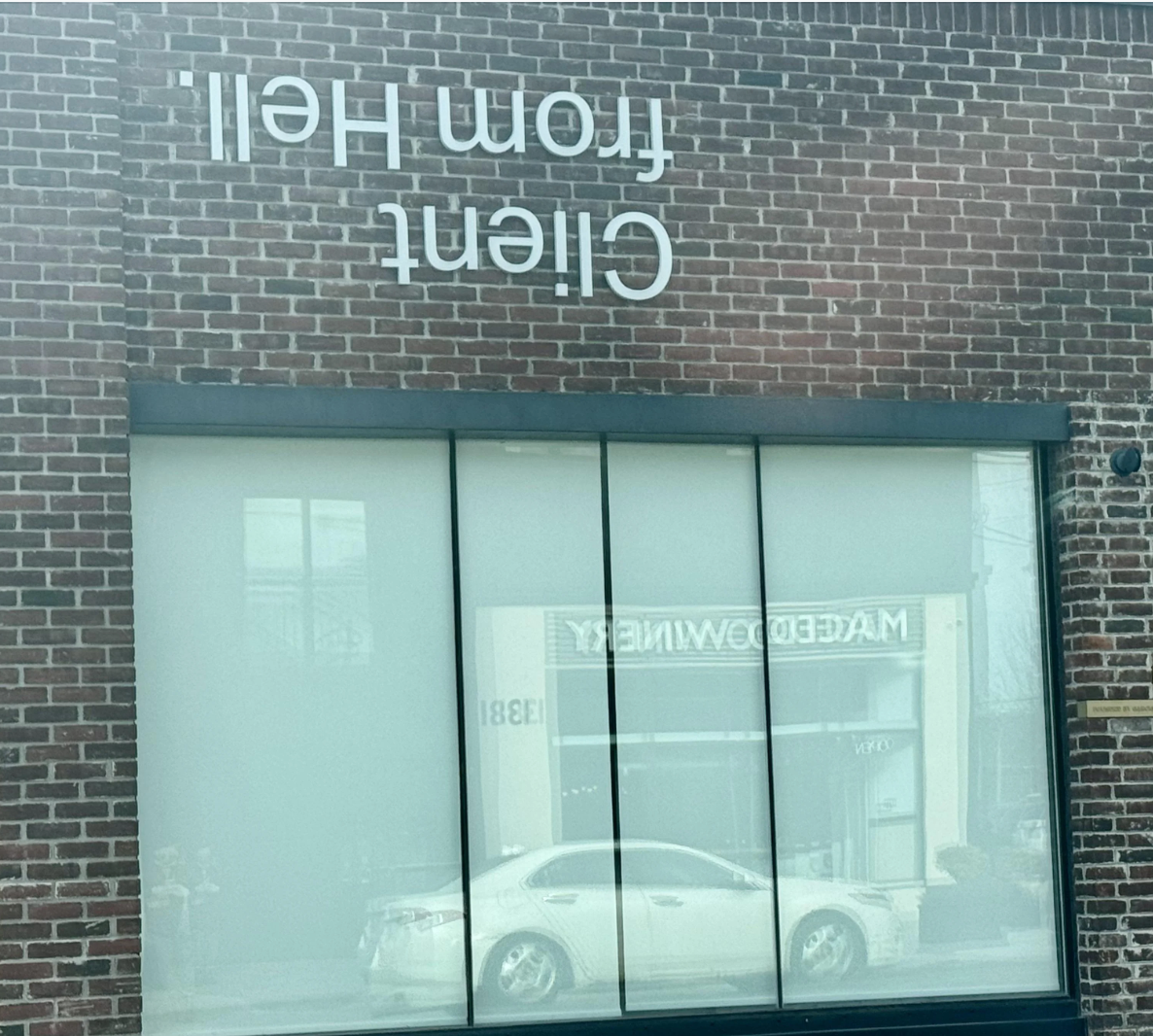 Brick wall with reversed text reading &quot;Client from Hell&quot; and a reflection of a white car and visible text &quot;MACEDO WINERY.&quot;