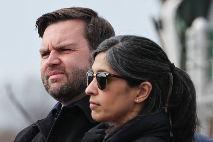 Two people outdoors, one wearing a dark coat, the other with sunglasses and earrings. Both are looking to the left