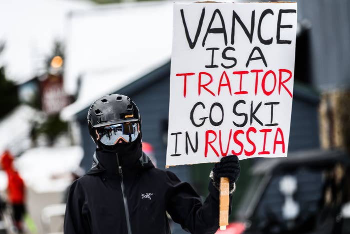 Person in ski gear holding a protest sign reading, &quot;Vance is a traitor, go ski in Russia,&quot; at a snowy location