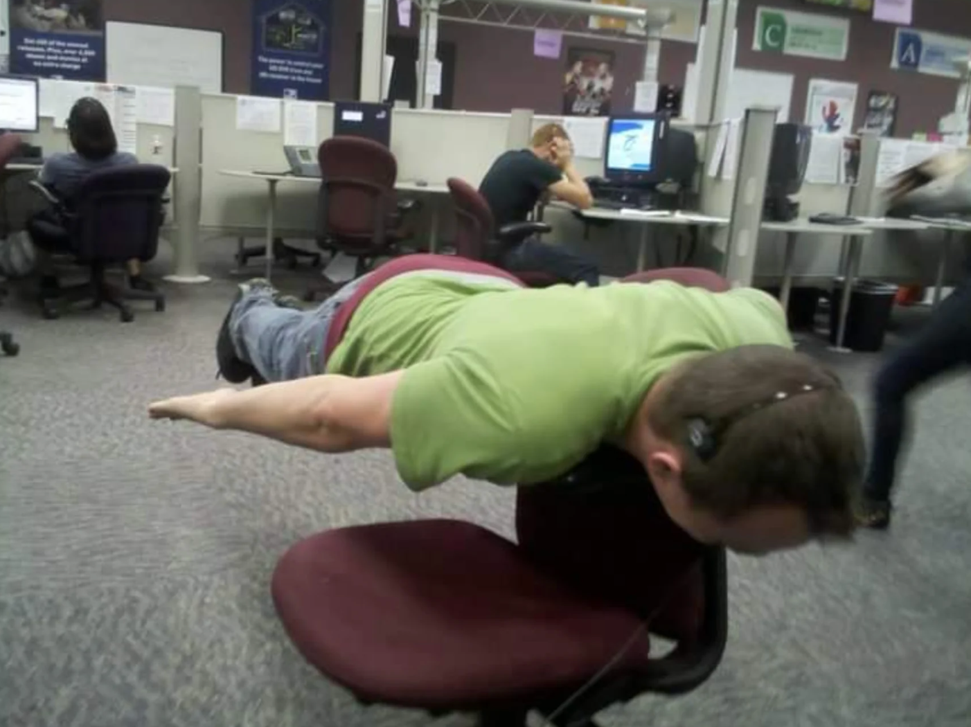 Person performs the planking meme on an office chair in a workplace with cubicles and seated employees in the background