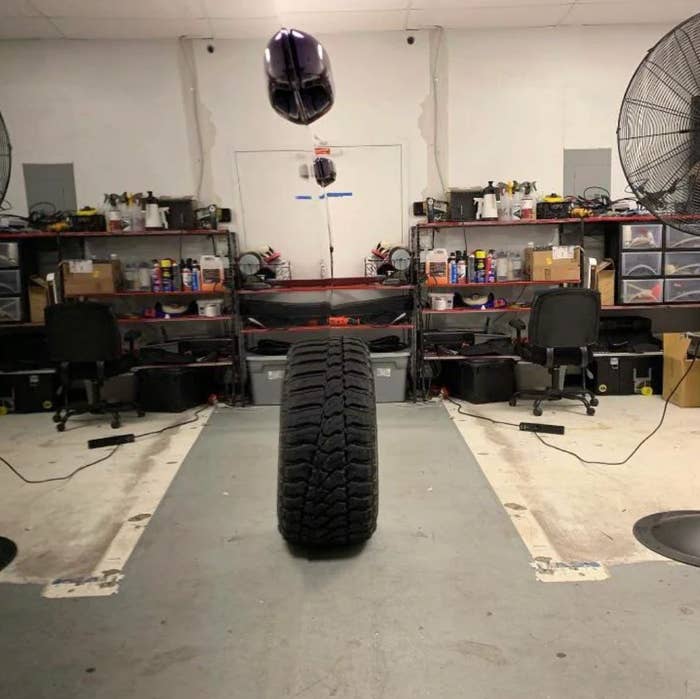 A tire is suspended mid-air in a workshop filled with shelves, tools, and two fans. Two chairs are placed near the shelves