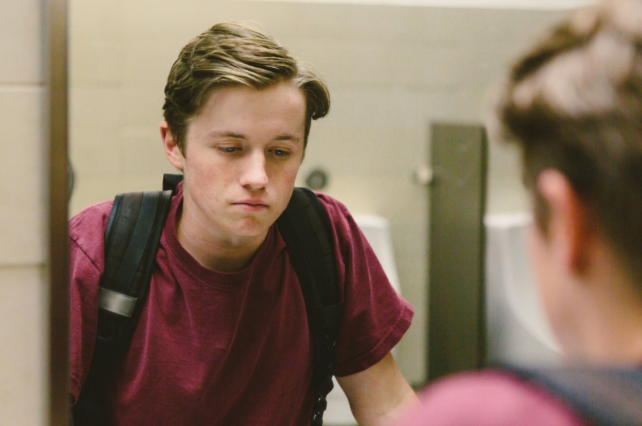 Teen wearing a t-shirt and backpack looks thoughtfully into a mirror in a restroom setting