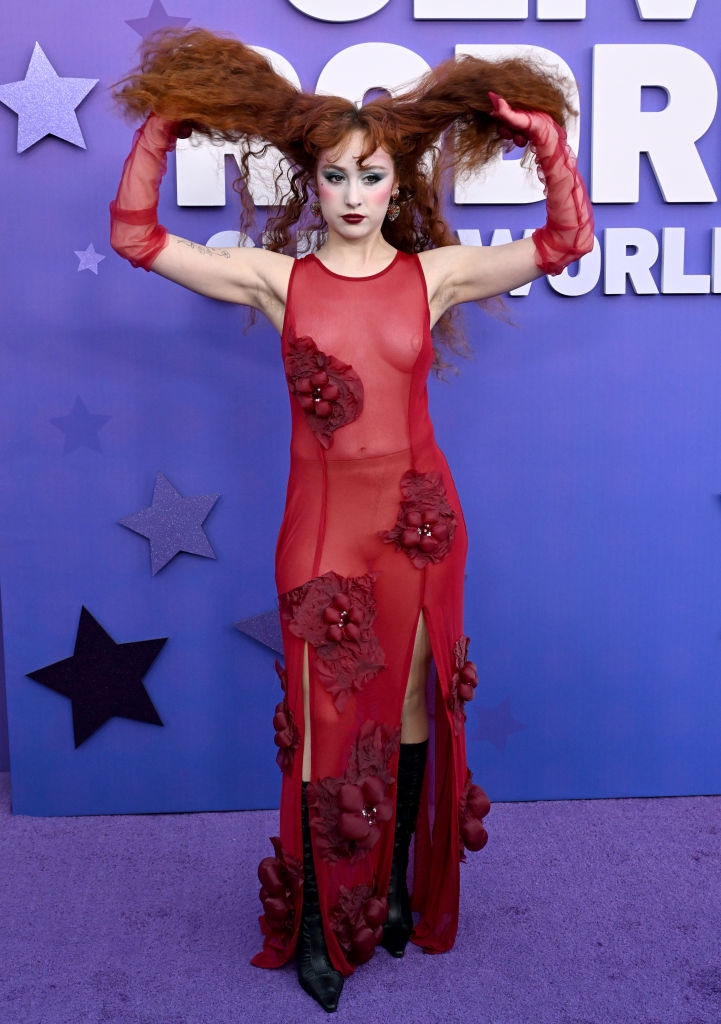Person in sheer, embellished dress with flower details poses playfully, lifting hair up, against a starry backdrop at a public event