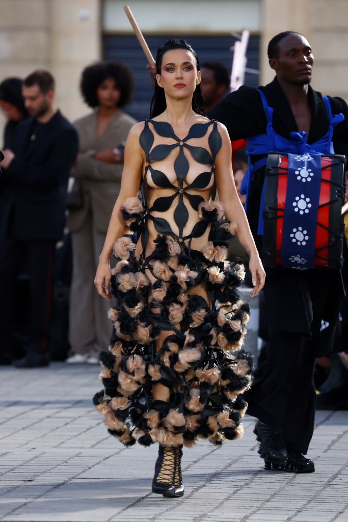 Person in unique, artistic dress with abstract patterns and ruffled details, walking in a public setting with people and performers in the background