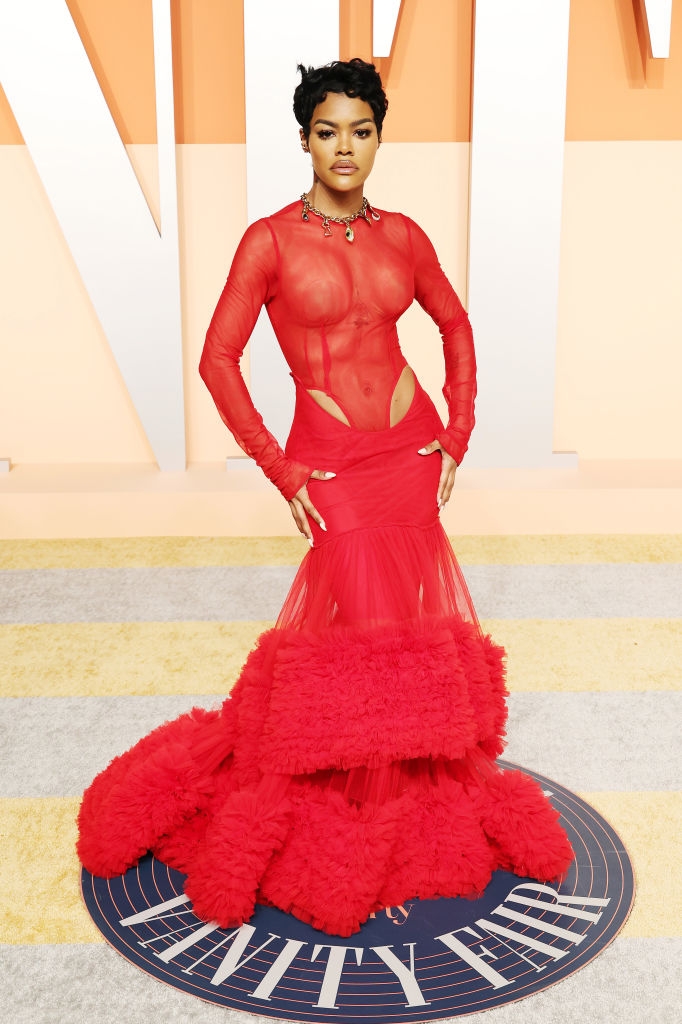 Person in a sheer, long-sleeved dress with dramatic cutouts and a voluminous textured skirt poses confidently on the red carpet