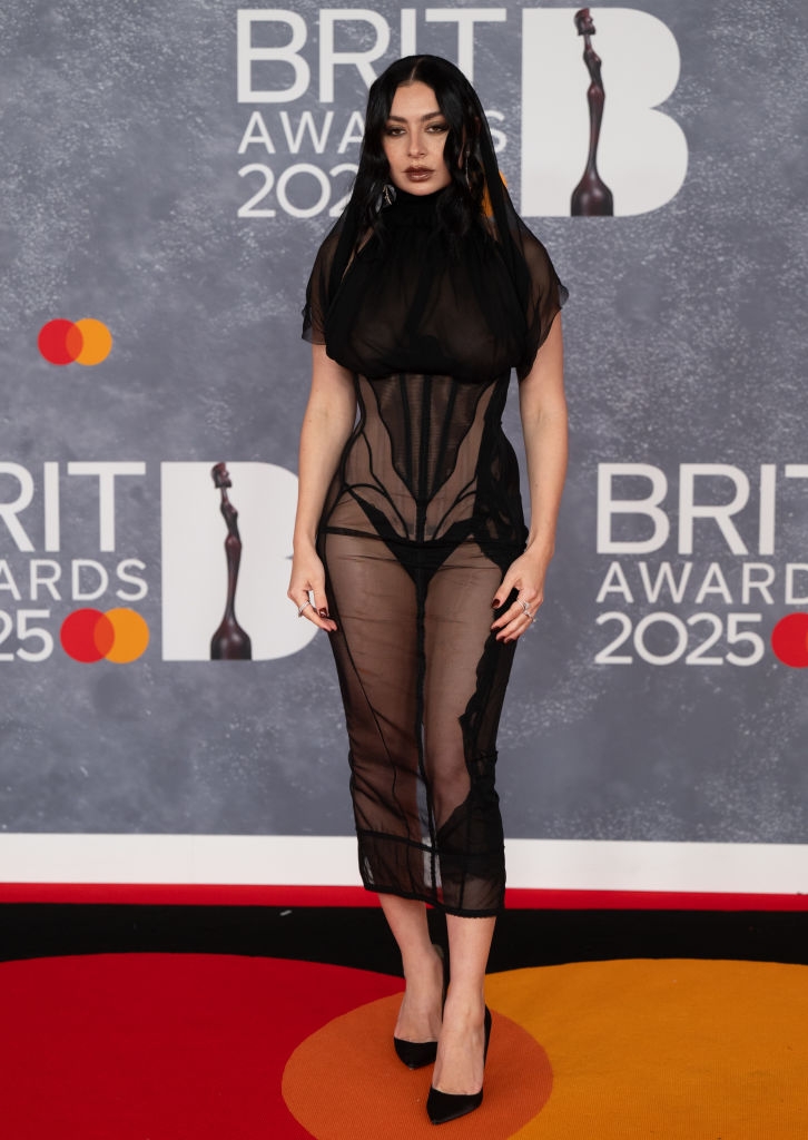 Person on a red carpet in a sheer, form-fitting dress with shoulder draping at the BRIT Awards 2025