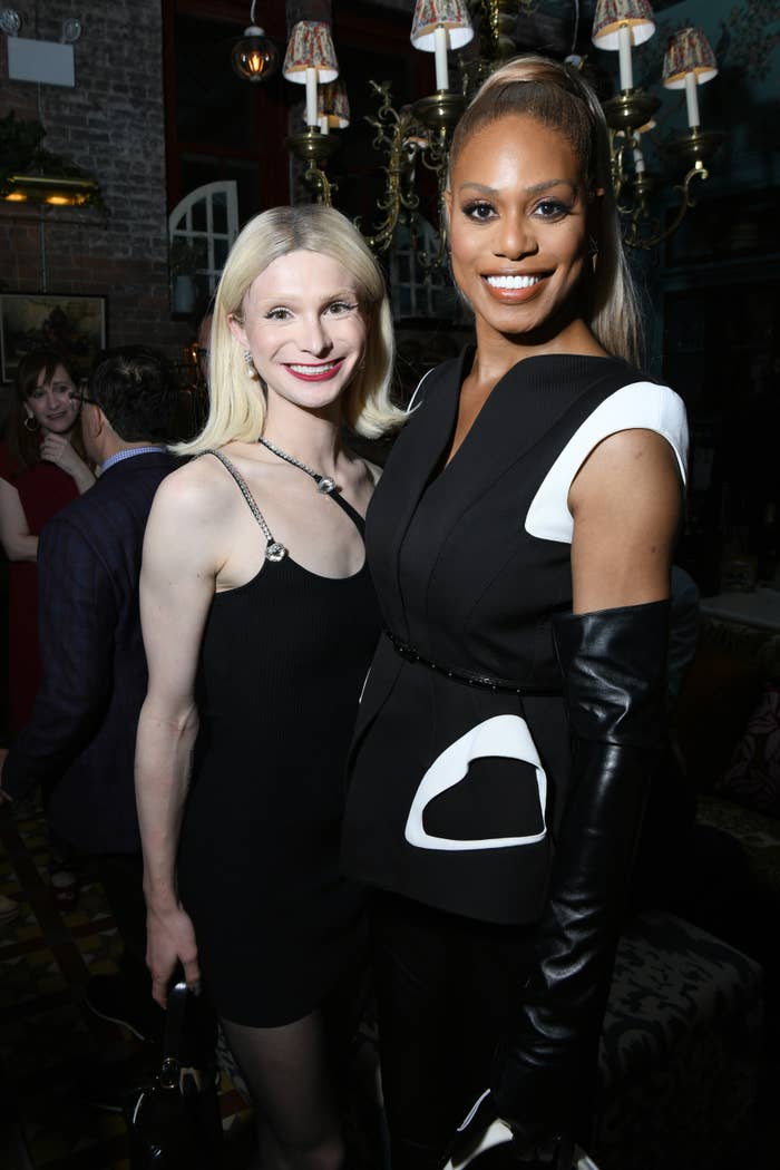 Dylan Mulvaney in a short dress with thin straps that cross over her right shoulder and tights with her hair flipped, standing next to Laverne Cox who&#x27;s wearing a sleeveless blazer style dress with tall leather gloves and a high ponytail