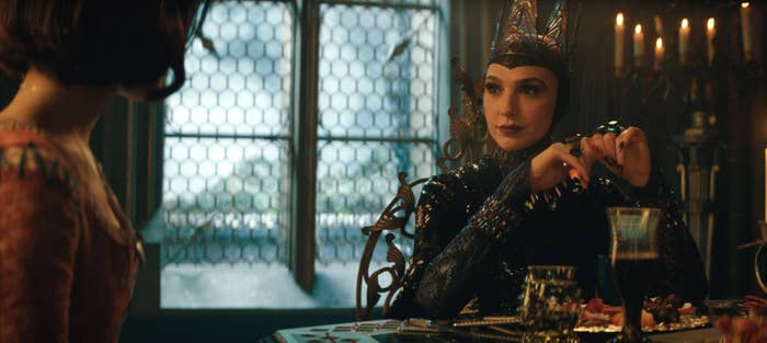 Gal Gadot and Rachel Zegler in an ornate room, one wearing an elaborate headpiece and detailed costume, sitting at a table with food and drink