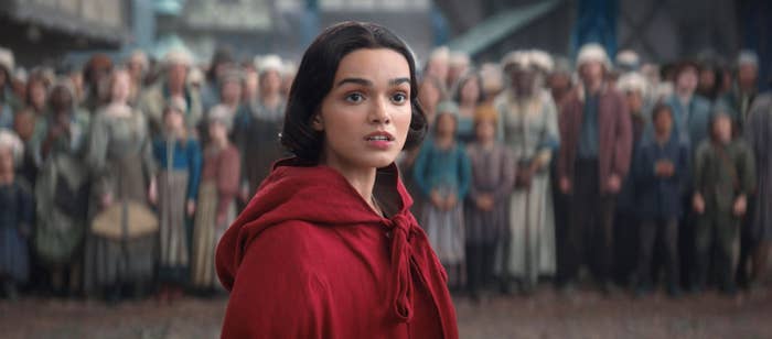 Rachel Zegler in a red cloak stands in a crowded scene, looking back with a concerned expression