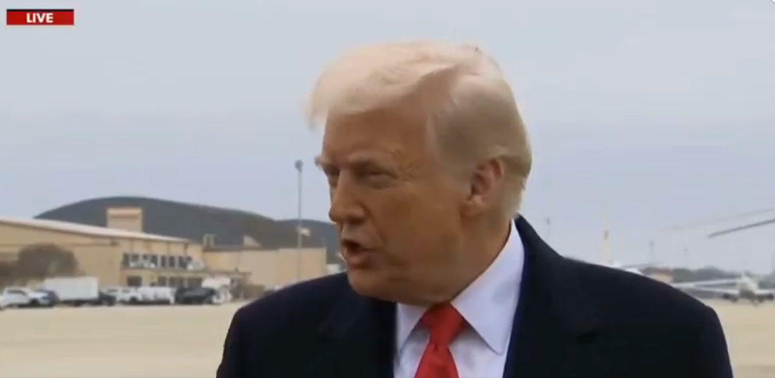 Trump in a suit stands on an airport tarmac, speaking