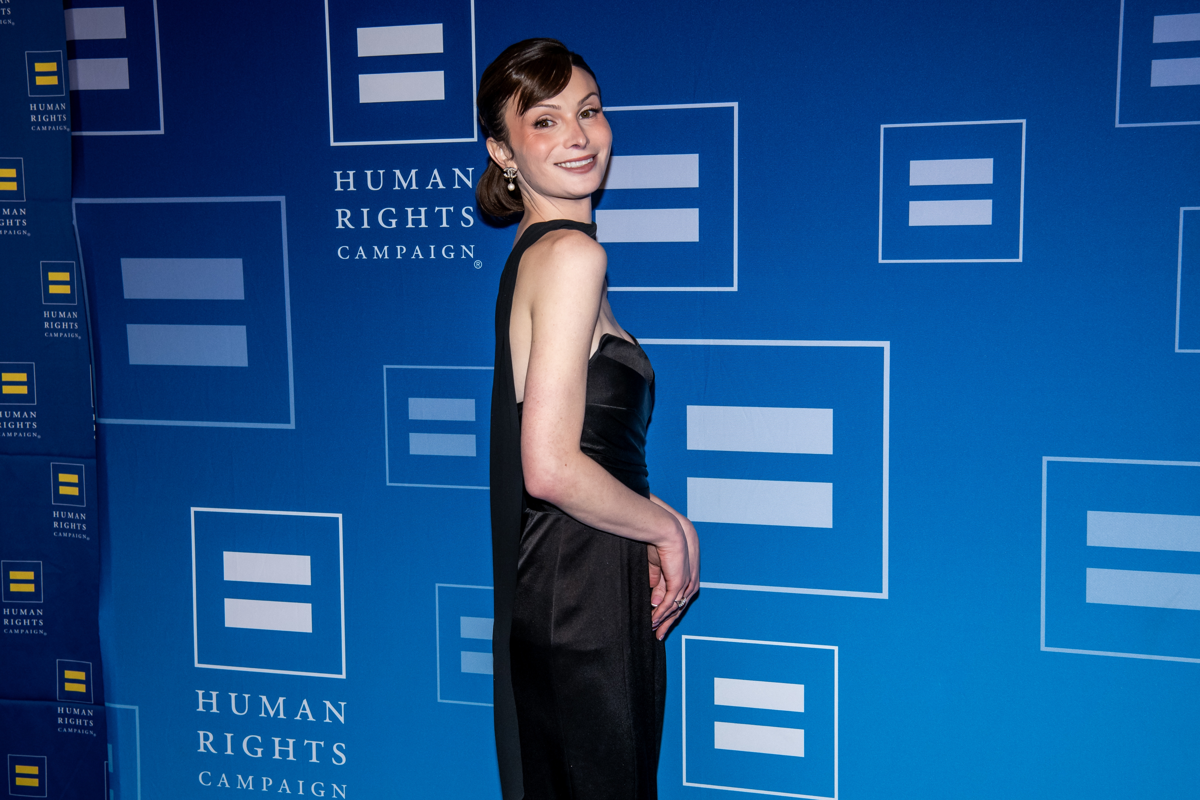 Dylan Mulvaney in a sleek gown at Human Rights Campaign event, standing against logo backdrop, smiling at the camera