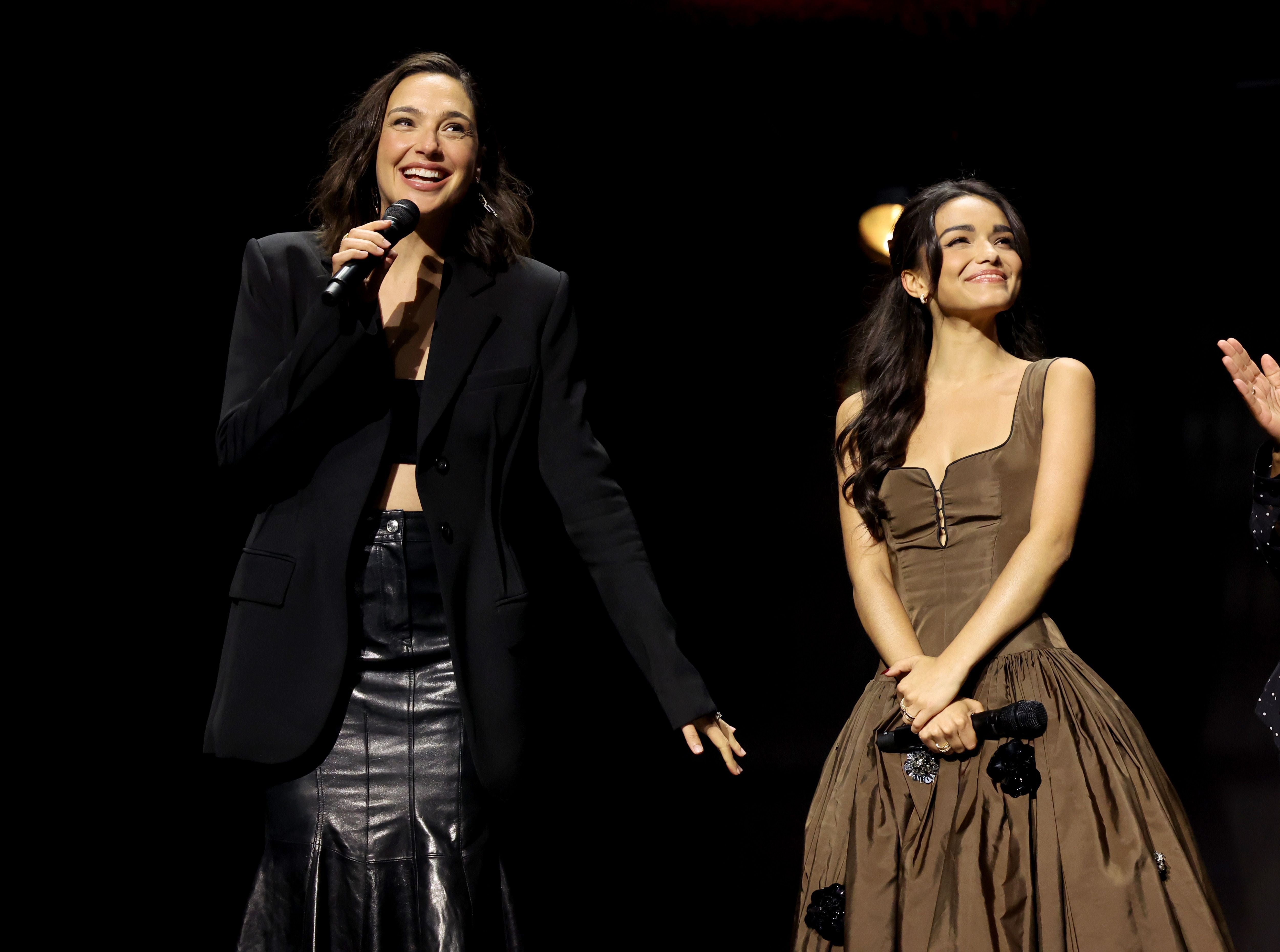 Gal Gadot and Rachel Zegler on stage, one in a blazer and skirt holding a mic, the other in a strapless dress smiling warmly