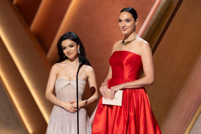Rachel Zegler and Gal Gadot at an event. One wears a strapless gown with delicate details; the other wears a bold strapless dress with a gold necklace, both smiling