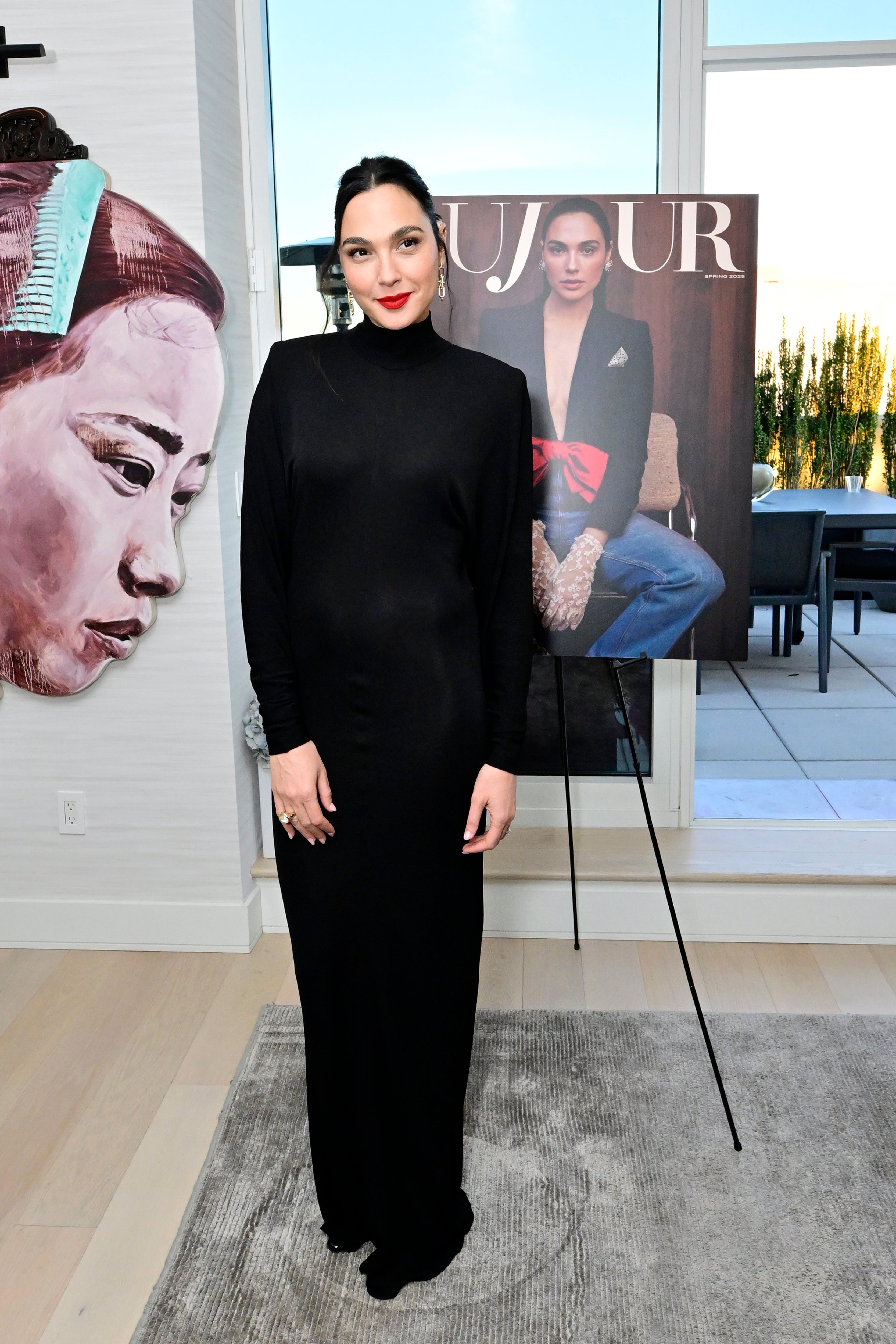 Gal Gadot in a long black dress poses next to a magazine cover featuring her. Art of a face is visible on the wall