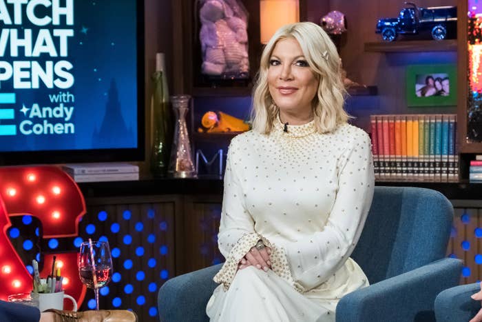 Tori Spelling seated in a TV studio, wearing a chic long-sleeved dress, watching an interview with books and decor visible in the background