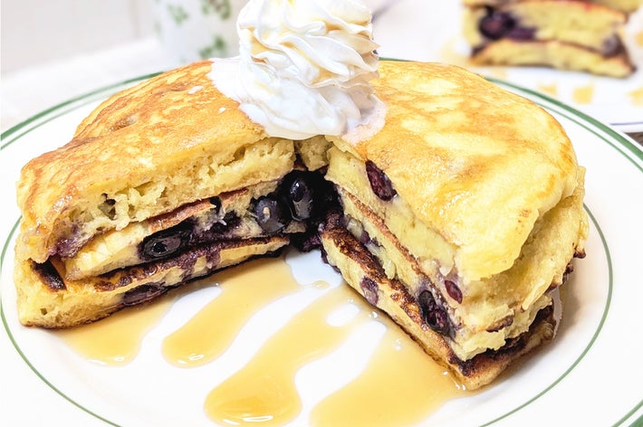 パンケーキにシロップとホイップクリームが載せられ、ブルーベリーが中に挟まれています。背景にカップが見える。
