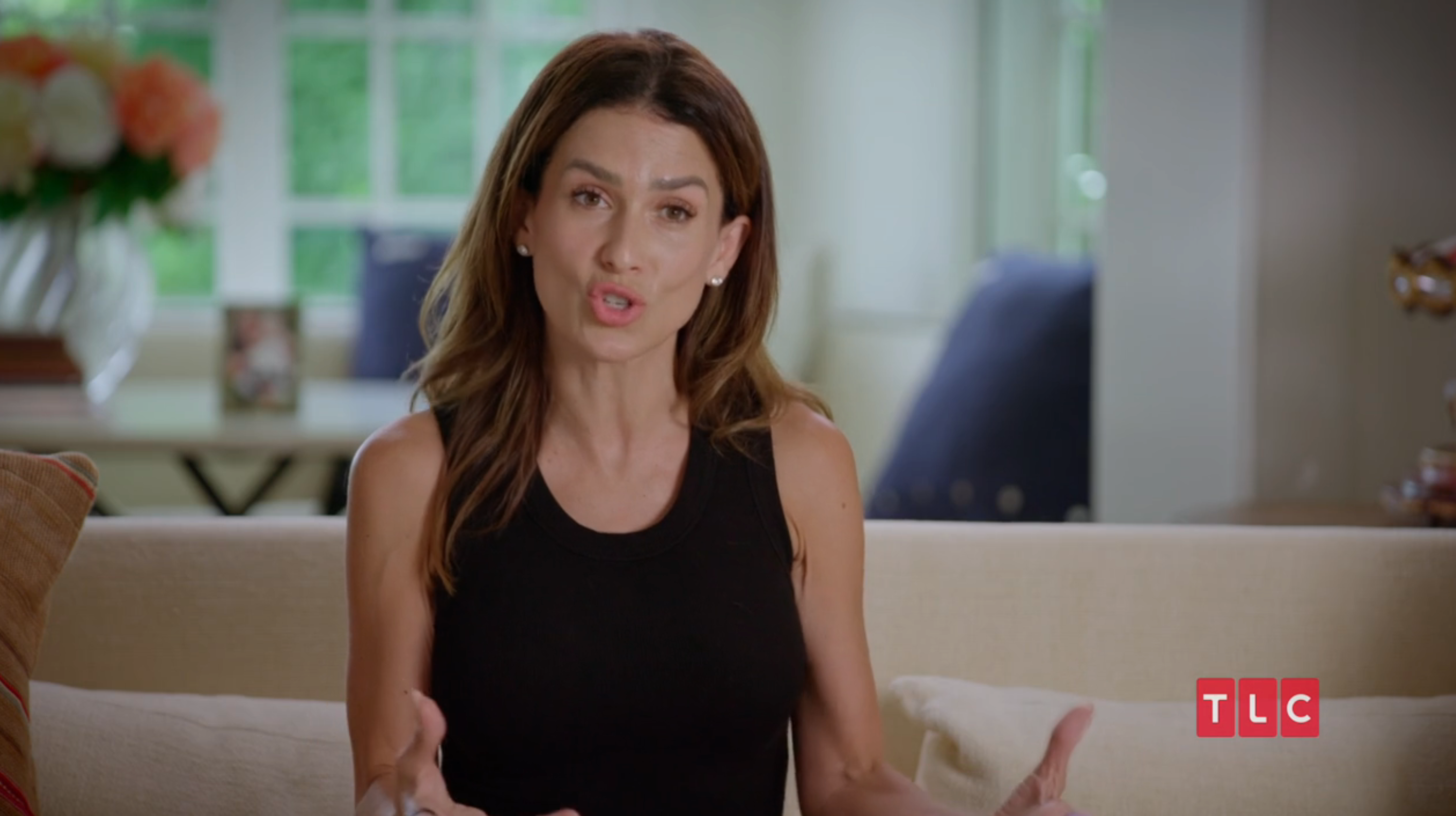 A person wearing a sleeveless top gestures while speaking on a sofa, appearing in a TLC program