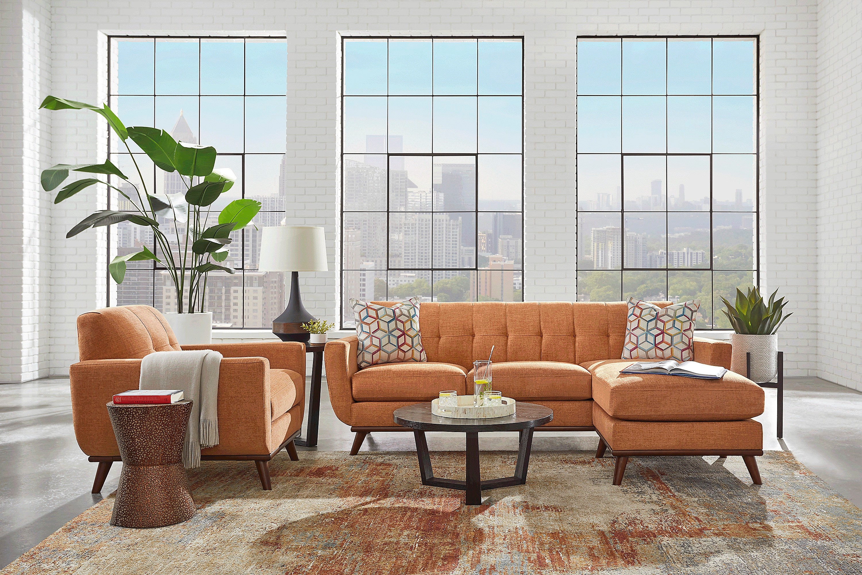 Modern living room with an orange sofa set, decorative pillows, a round coffee table, and a tall potted plant near large windows