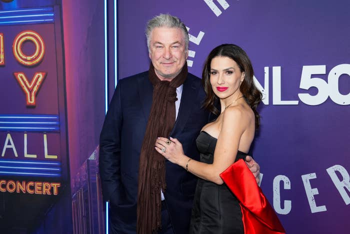 A man in a dark coat and scarf poses with a woman in an elegant black dress with a red bow on the red carpet at an event