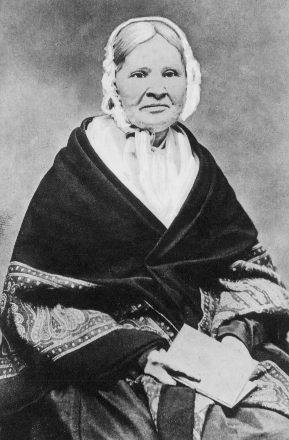 An elderly woman in a historical outfit and lace bonnet sits holding a book. She wears a shawl draped over her shoulders