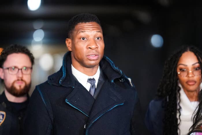 I don’t know who this is, but the image shows a man in a tailored coat over a suit, flanked by two people, possibly in a formal or public setting