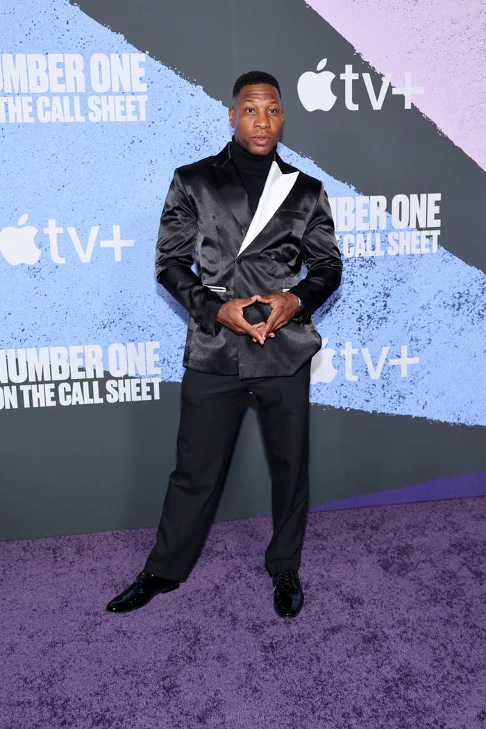 Person on a red carpet wearing a stylish black suit with a white accent shirt, posing in front of a backdrop featuring the Apple TV+ logo