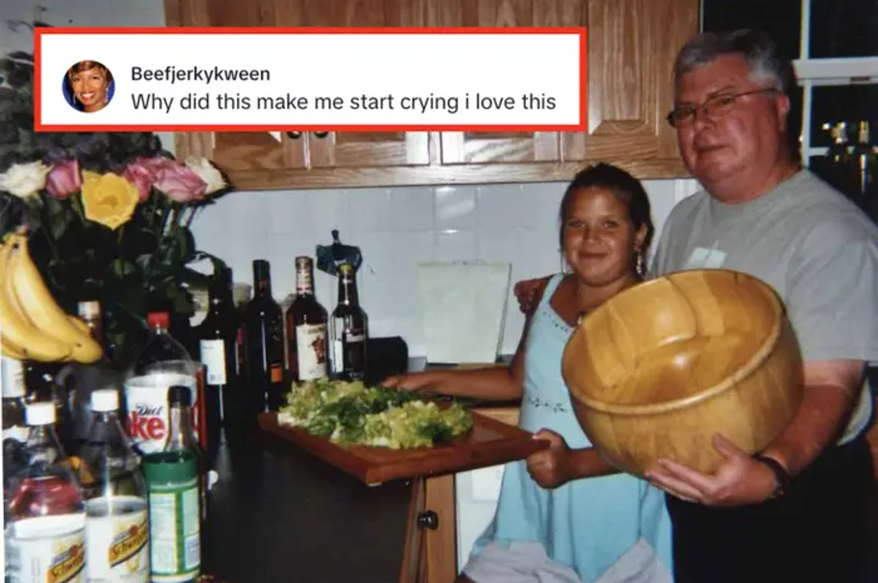 Two people smiling in a kitchen holding a salad in a large wooden bowl and serving platter, surrounded by bottles and flowers