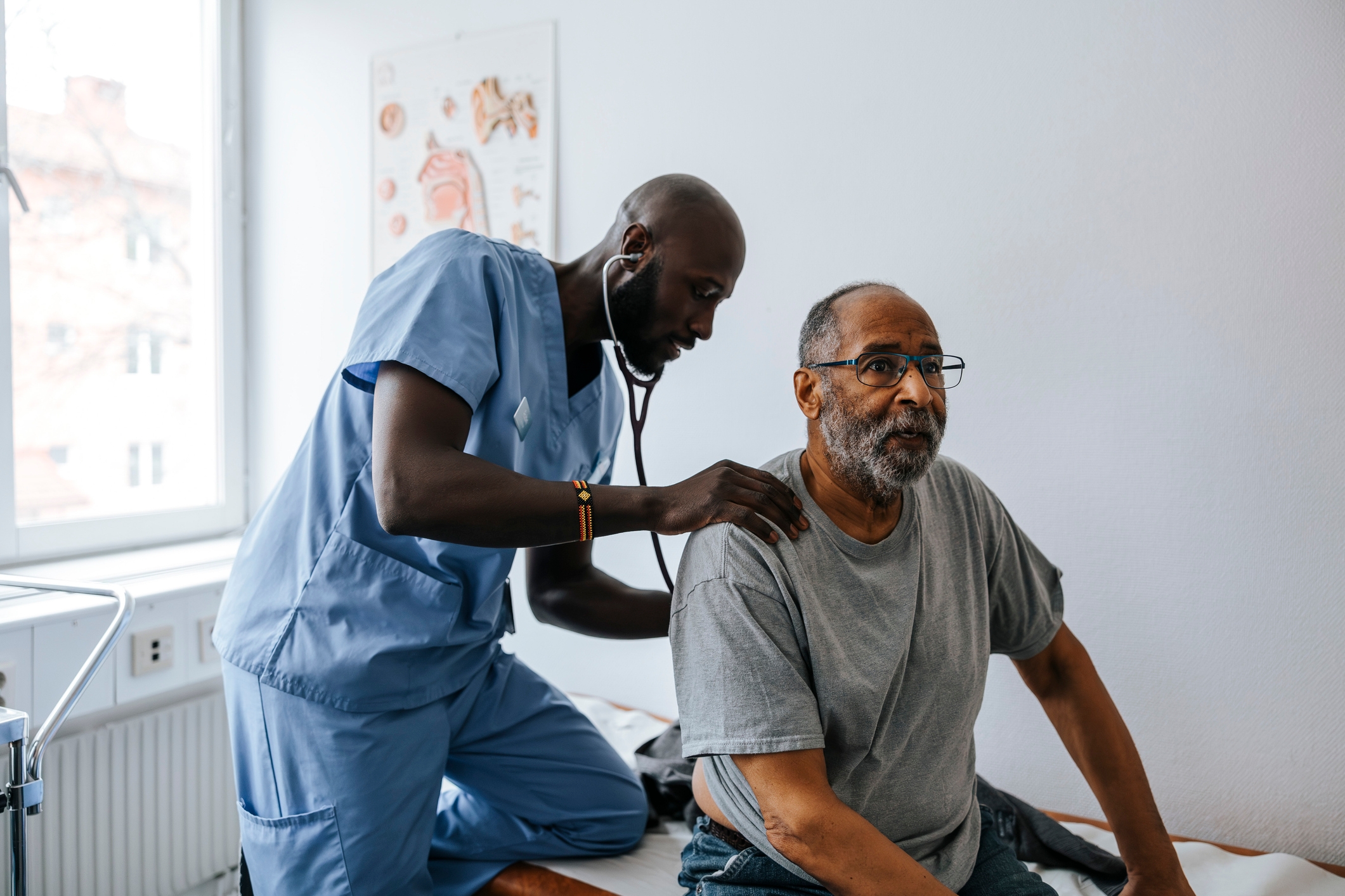 Doktor i Scrubs bruker et stetoskop på en sittende manns tilbake på et legekontor, med fokus på helse og velvære