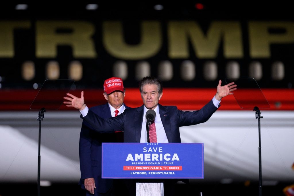 To menn på en rally -scene; Man snakker på et podium med & quot; Save America & quot; Tekst, den andre har på seg en merkevarehette, som står bak et stort fly merket & quot; Trump. & quot;