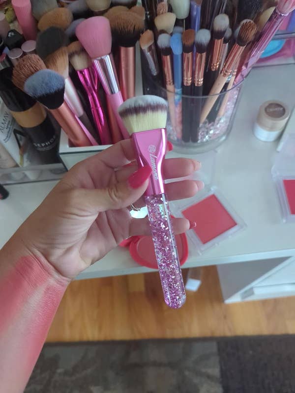 Hand holding a makeup brush with sparkling, transparent handle. Various makeup brushes in a holder are in the background