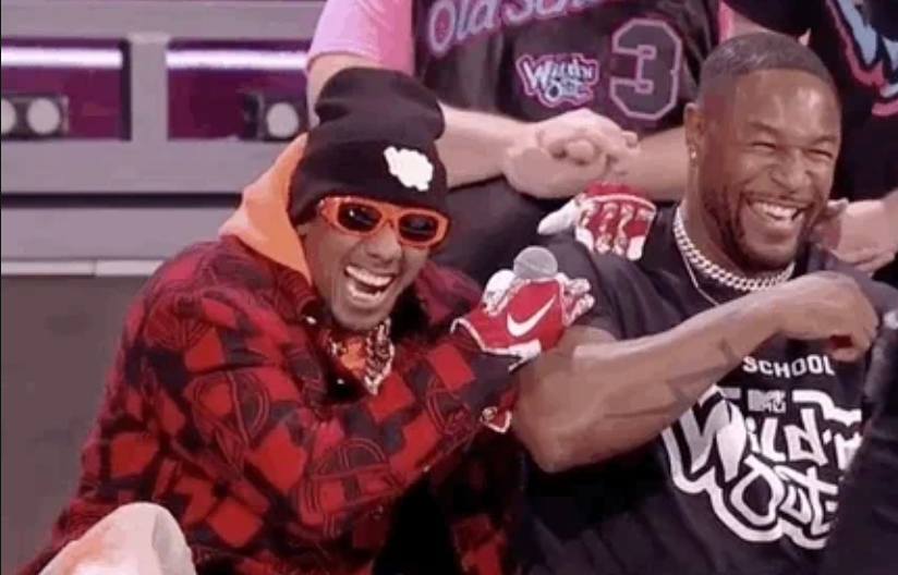 Two people on a comedy show stage are laughing together. One wears a patterned jacket and sunglasses, while the other has a logo t-shirt