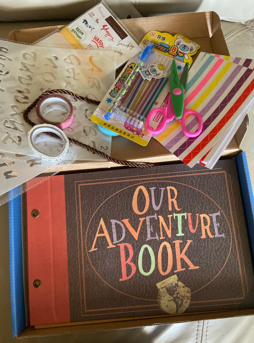 Scrapbooking kit with a book labeled "Our Adventure Book," stickers, colored pens, scissors, and washi tape on a tray