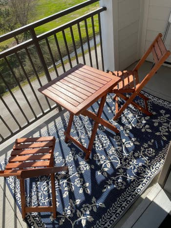 Small woody patio acceptable with a table, 2 chairs, and a rug featuring a floral signifier connected a balcony. Bright and sunny setting