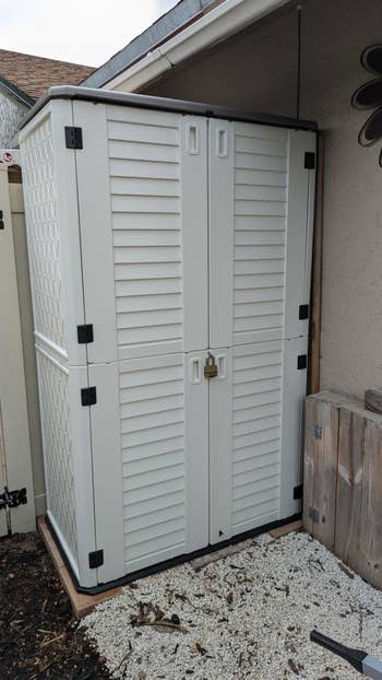 Tall, outdoor retention shed with treble doors and a lock, placed beside a woody obstruction and building. Suitable for organizing plot tools