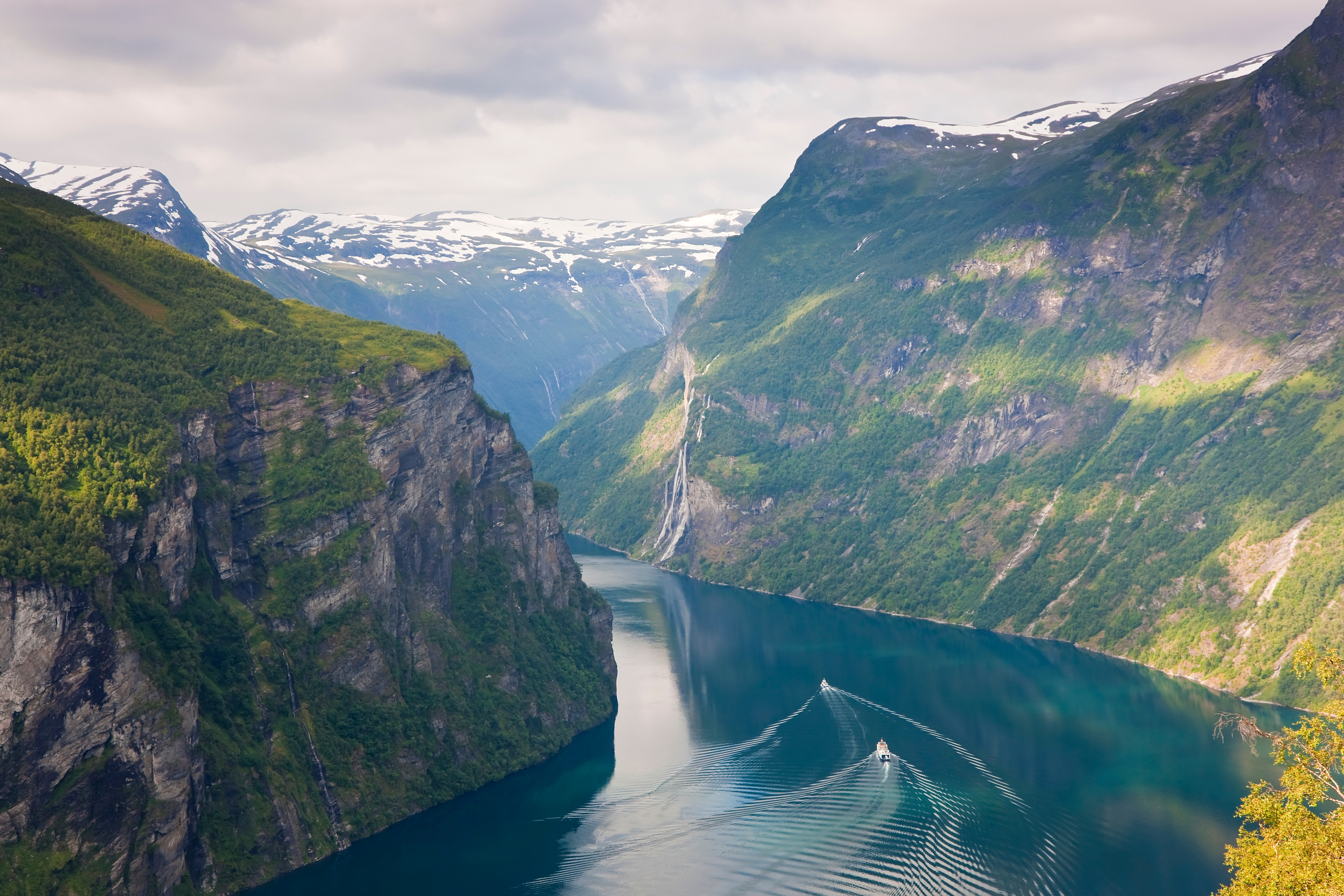 Fjellrike landskap med en smal fjord, bratte klipper og en liten båt som etterlater krusninger i vannet. Snødekte topper er synlige i det fjerne