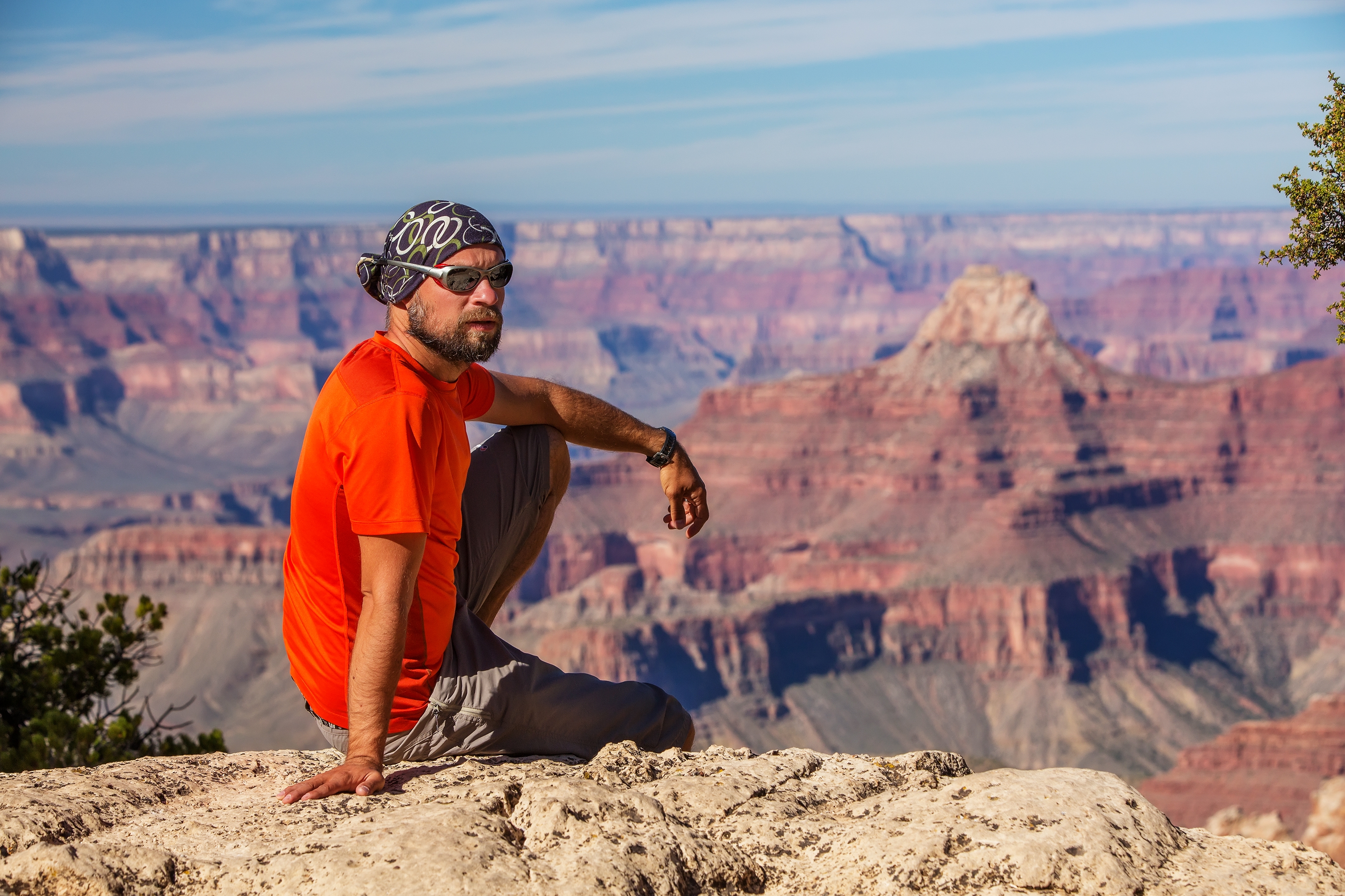 Mannen som sitter på en stein avsats med utsikt over et stort canyonlandskap. Han har på seg uformelle uteklær og solbriller
