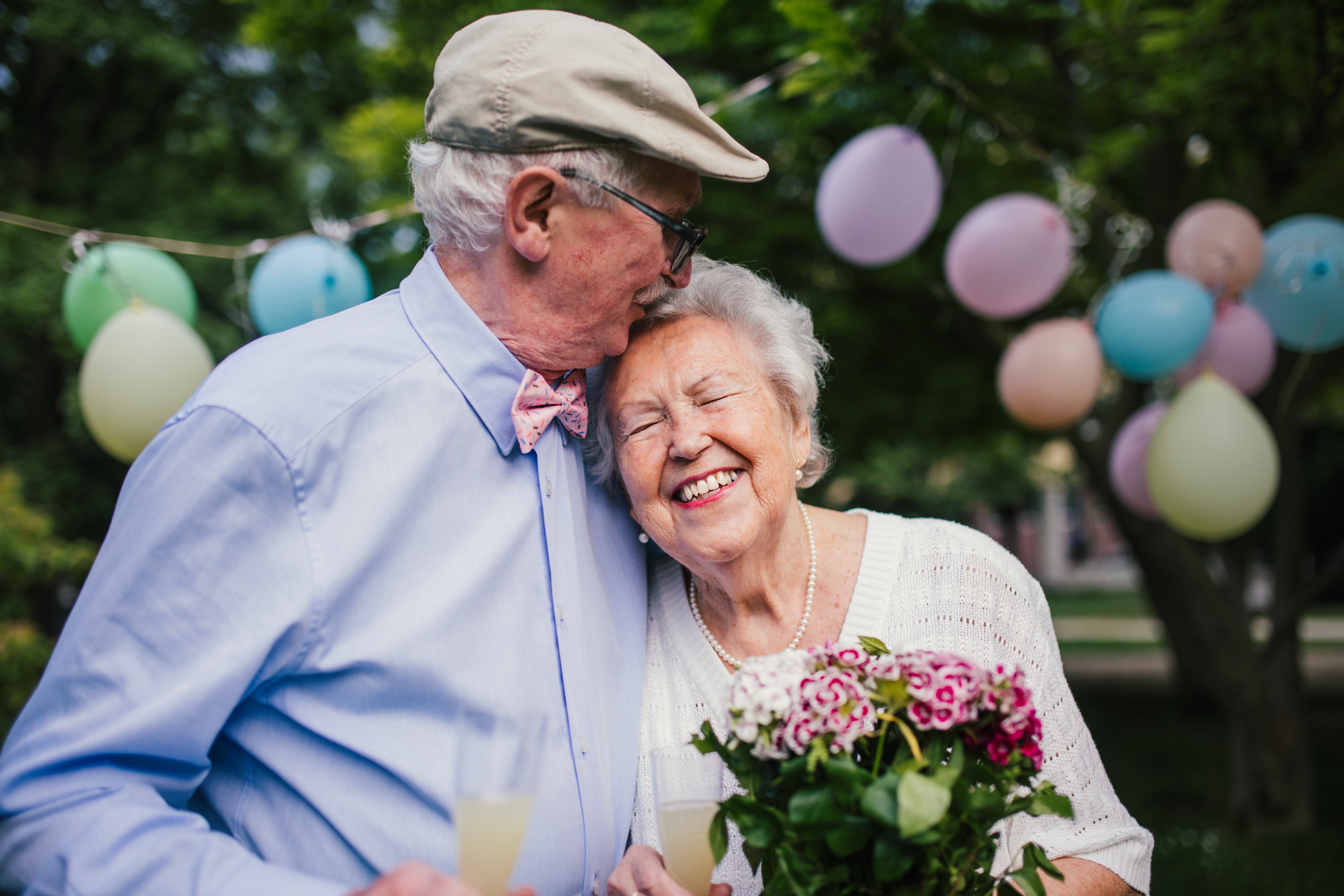 Eldre par feirer med blomster og ballonger, mann kyss kvinnens hode mens hun holder bukett, begge smiler lykkelig