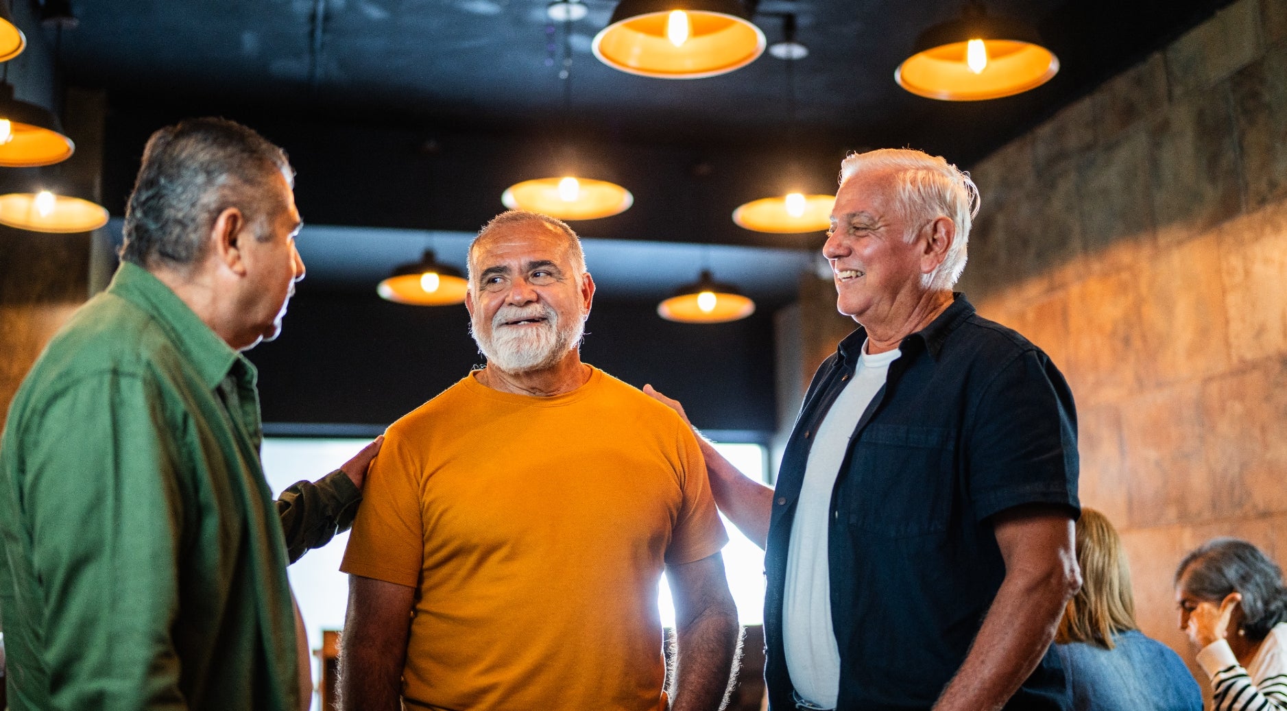 Tre eldre menn i uformelle klær chatter og smiler i en varmt opplyst restaurant eller kafé